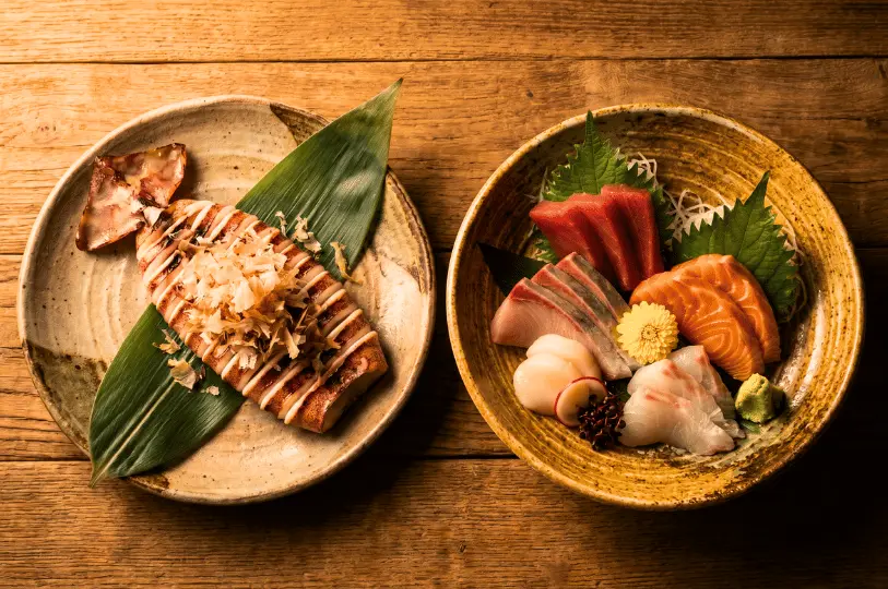 Two beautifully plated Japanese dishes on a rustic wooden table: a whole grilled squid topped with bonito flakes resting on a bamboo leaf, and a colorful sashimi platter featuring fresh cuts of tuna, salmon, and scallops.