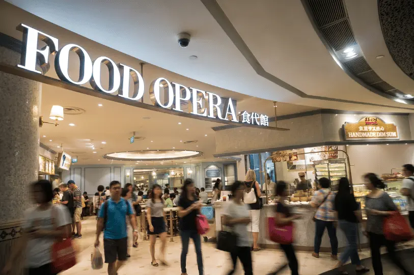 A lively, busy scene at the brightly lit entrance of Food Opera in ION Orchard, with numerous motion-blurred shoppers walking past a traditional 
