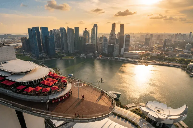 A sweeping high-angle view from the Marina Bay Sands observation deck at sunset, looking down at a lower dining terrace with red umbrellas, the bay waters, and the dense Singapore cityscape.