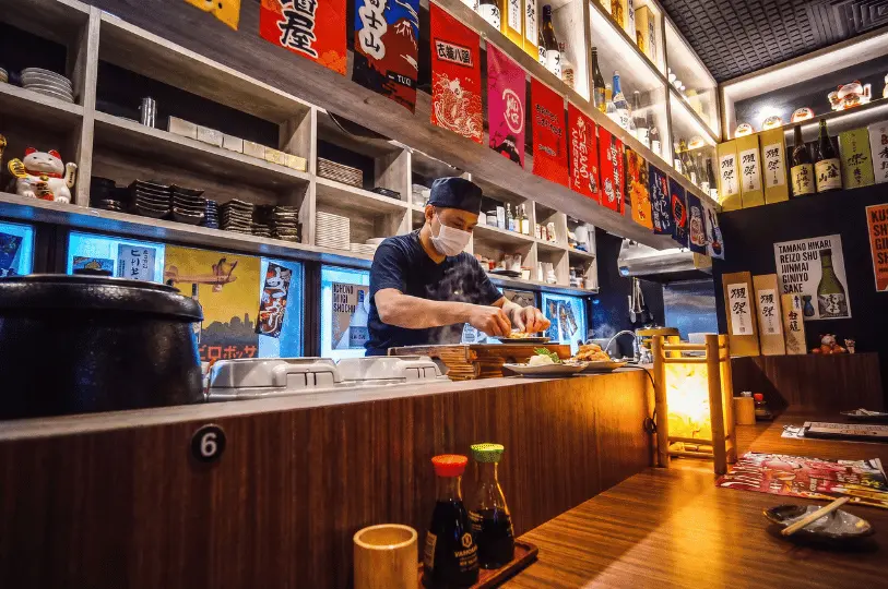 A cook wearing a face mask and cap preparing dishes behind a warm wooden counter bar at Izakaya Hikari, surrounded by shelves of sake bottles, decorative banners, and soy sauce dispensers.