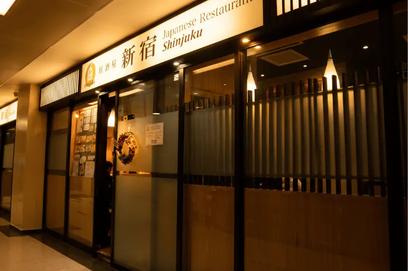 The exterior of Shinjuku Japanese Restaurant at Cuppage Plaza, featuring bright overhead signage and warm interior light glowing behind slatted wooden screens and glass doors.