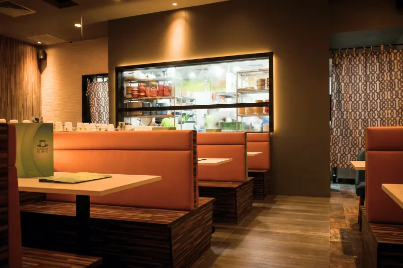 The clean, modern dining area of a casual sushi restaurant featuring cozy orange booth seats, warm ambient lighting, and a window into the bright kitchen area.