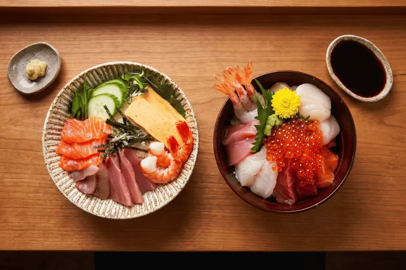 A top-down view of two Japanese seafood rice bowls on a light wooden table. The left bowl features neatly arranged sliced sashimi, cooked shrimp, and tamago, while the right bowl is piled high with raw sweet shrimp, scallops, sashimi, and salmon roe.