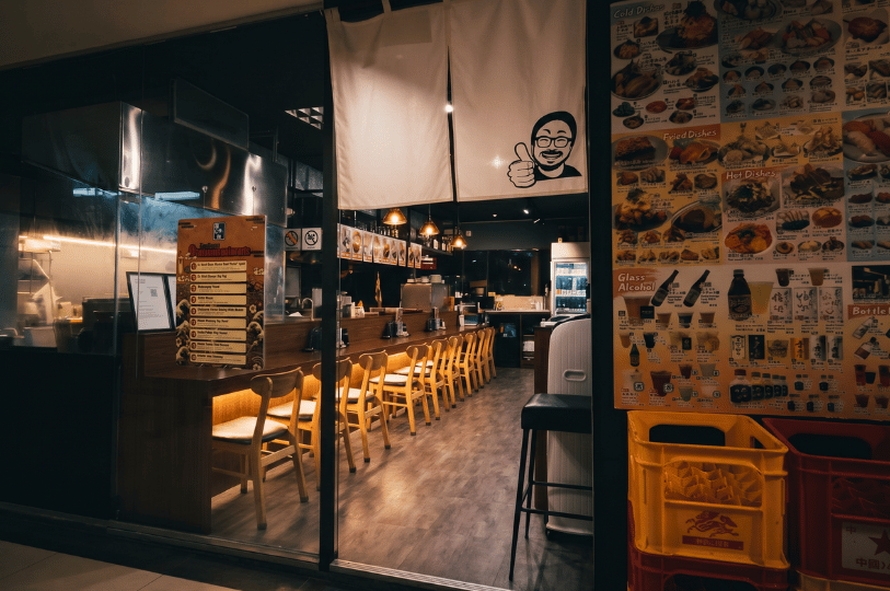 The exterior storefront of a warmly lit Japanese-style restaurant, featuring a massive, floor-to-ceiling menu on the right wall densely packed with dozens of food photos and drink options. Noren curtains hang above the glass entrance, revealing an empty, long wooden dining counter lined with chairs inside.