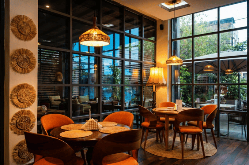 A warm and inviting restaurant interior featuring round wooden tables with orange-upholstered chairs, illuminated by glowing rattan pendant lights and a vintage fringed floor lamp. Large glass grid windows reveal a leafy outdoor patio, while a white wall pillar is adorned with a vertical column of woven straw sunburst decorations, setting a cozy and earthy dining mood.