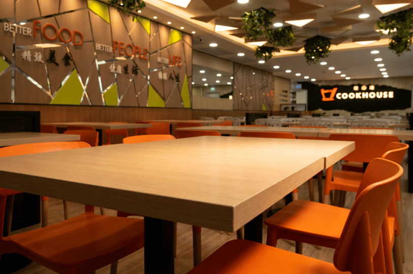 A close-up, low-angle view focusing on an empty, light-wood laminate table in a bright, modern food court, surrounded by vibrant orange chairs. In the softly blurred background, an illuminated sign reads "Cookhouse" alongside a geometric wall graphic stating "Better Food, Better People, Better Life", highlighting the everyday communal nature of the space.
