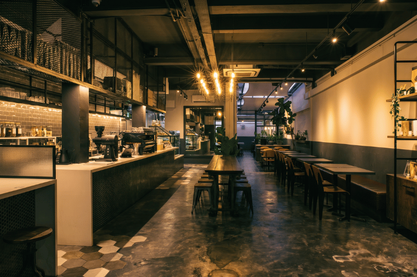 An empty, industrial-chic cafe interior featuring a long coffee bar with espresso machines on the left, warm hanging pendant lights, and a mix of communal and private wooden seating across a polished concrete floor, illustrating the upscale dining atmosphere found in modern shopping spaces.