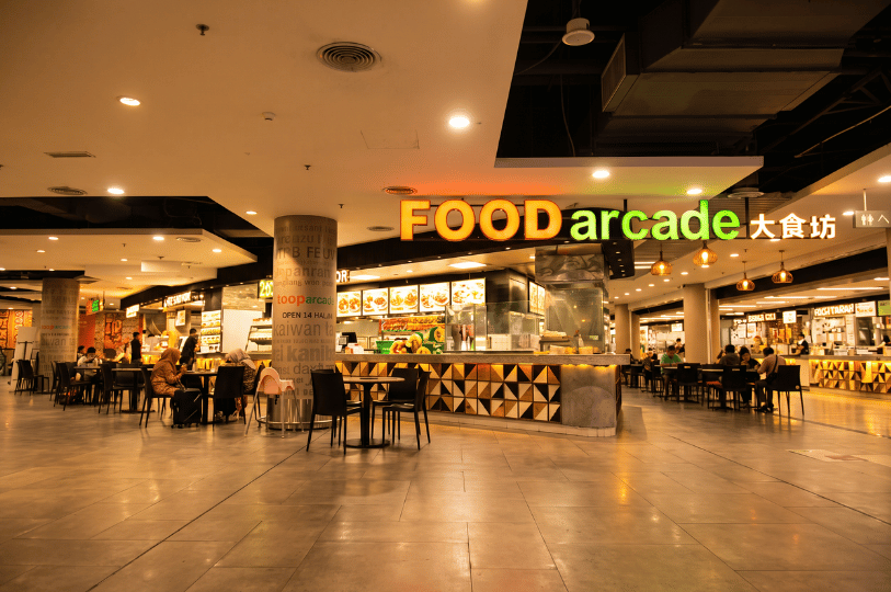 A wide, brightly lit view of the FOOD arcade in a shopping mall. Patrons are seated at scattered black tables, surrounded by a diverse array of food stalls with illuminated menu boards, illustrating the overwhelming variety of dining choices.