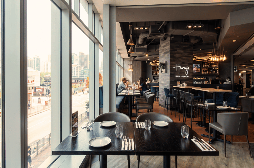A modern restaurant interior featuring an empty, set dining table positioned directly beside large floor-to-ceiling windows, offering a bright view of a bustling city street and illustrating the appeal of natural light and window seating.