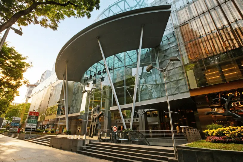 Low-angle wide shot of the grand, curved glass entrance canopy of Paragon Shopping Centre, framed by lush trees and high-end luxury storefronts.