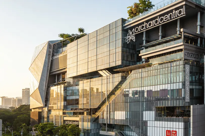 The towering, multi-tiered glass exterior of Orchard Central mall reflecting the warm golden hour sun, highlighting its complex modern architecture and outdoor escalators.