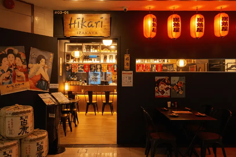 The entrance to Izakaya Hikari (unit #03-01) at Fortune Centre, glowing with red paper lanterns, traditional sake barrels stacked outside, vintage posters, and a view into the warm interior.