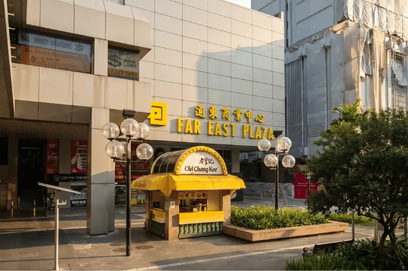 Street-level view of the Far East Plaza entrance, featuring its blocky grey facade and a small, bright yellow Old Chang Kee food kiosk in the paved plaza.