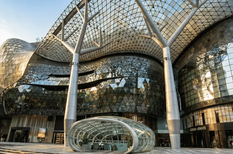 The futuristic glass and steel facade of ION Orchard shopping mall, featuring massive tree-like structural pillars and a curved glass canopy over the MRT entrance.