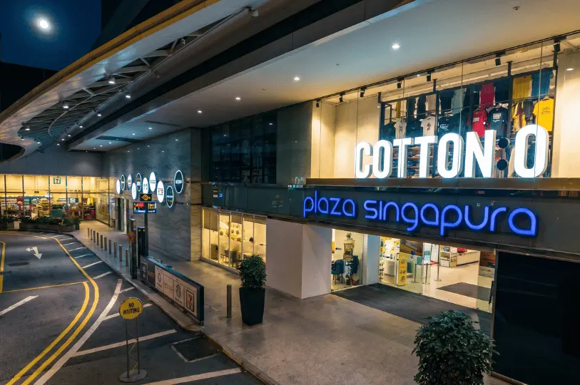 Nighttime view of the Plaza Singapura mall entrance drop-off point, showing the illuminated blue mall signage, bright storefronts like Cotton On, and a quiet driveway under streetlights.