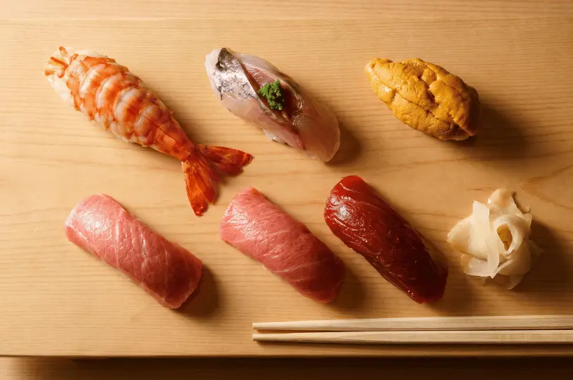 An overhead shot of six pieces of premium nigiri sushi—including otoro, akami, ebi, and uni—artfully arranged on a light wooden platter with pickled ginger and chopsticks.