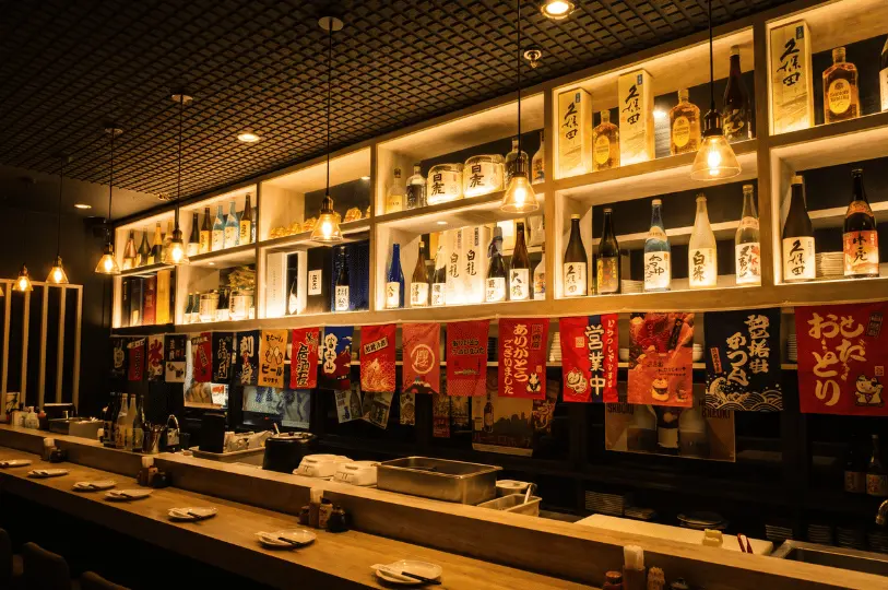 A long wooden bar counter illuminated by hanging filament light bulbs, lined with backlit glass shelves filled with sake bottles and colorful hanging Japanese flag banners.