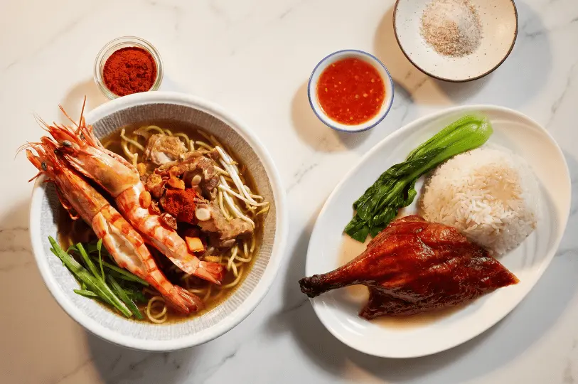 Top-down view of classic hawker dishes served on a white marble table: a hearty bowl of prawn and pork rib noodle soup alongside a plate of glossy roast duck with rice and greens.