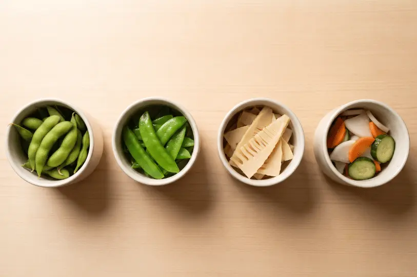 Four small white bowls arranged in a straight line on a light wooden surface, containing Japanese side dishes: edamame, snap peas, sliced bamboo shoots, and pickled vegetables.