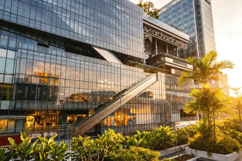 A wide exterior shot of the modern Orchard Central shopping mall in Singapore during sunset, featuring a multi-story glass facade, escalators, and tropical landscaping in the foreground.