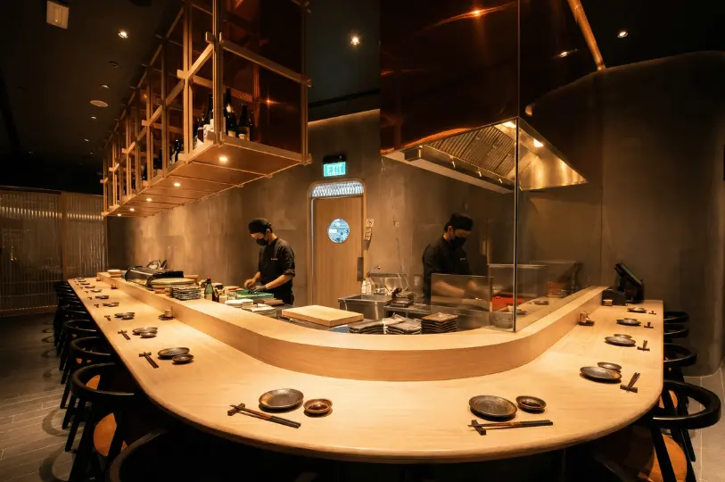 A sleek, modern restaurant interior showing a curved light-wood sushi counter, dark walls, suspended copper-toned shelving, and two chefs in black uniforms preparing food.