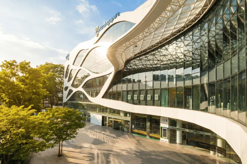 Daylight exterior view of the iconic Plaza Singapura shopping mall in Singapore, highlighting its futuristic white wave-like facade and extensive glass windows surrounded by lush green trees.
