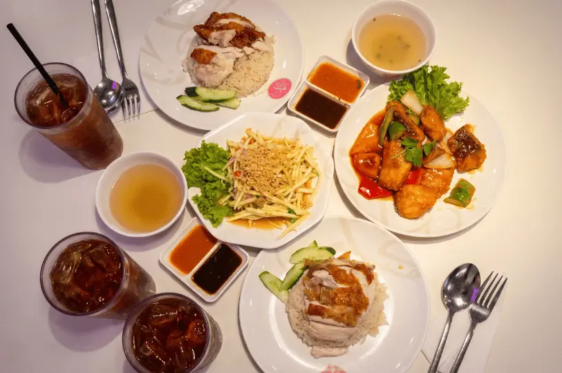 Overhead flat lay view of a diverse food court spread on a white table, including local chicken rice, sweet and sour stir-fried fish, mango salad, side soups, chili sauces, and large glasses of iced tea.