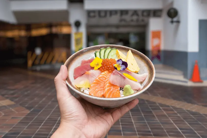A hand holding a vibrant bowl of kaisen don topped with thick slices of salmon, tuna, yellowtail, tamago, salmon roe, and edible flowers, with the blurred entrance of Cuppage Plaza in the background.