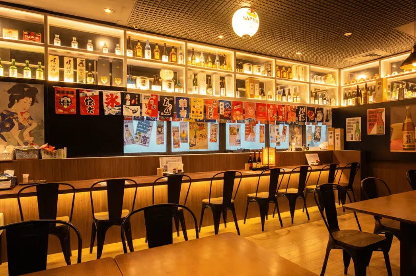The cozy dining interior of Izakaya Hikari showing wooden tables and counter seating under a modern dark ceiling, flanked by warm backlit shelves filled with sake bottles and vintage Japanese art posters.