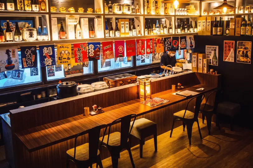 An intimate wooden counter setup inside Izakaya Hikari, with black metal chairs lined up before a illuminated back bar filled with sake bottles, decorative Japanese flags, and warm pendant lighting.