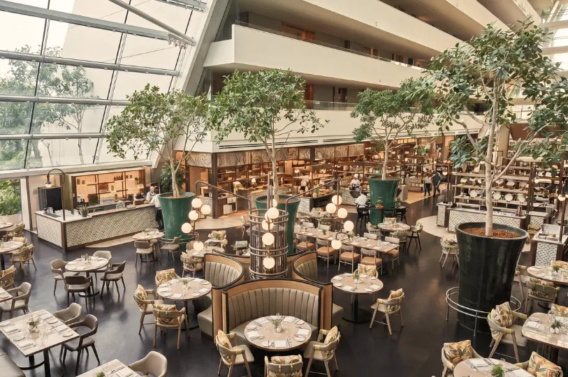 A spacious indoor atrium restaurant featuring high glass ceilings, large potted indoor trees, and multiple round dining booths arranged around central buffet stations.