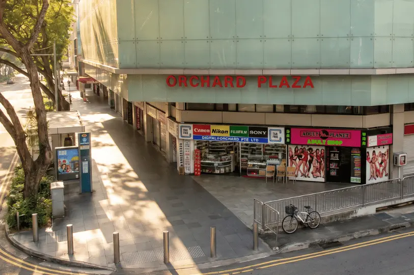 High-angle corner view of Orchard Plaza showing its pale green glass upper facade and street-level retail shops, including a camera store and an adult shop, next to an empty sidewalk.