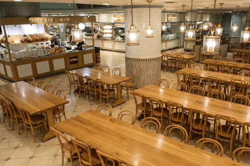 Wide shot of the Food Opera dining hall showing a juice bar and various stalls surrounding a spacious, empty seating area furnished with long wooden communal tables and vintage pendant lighting.
