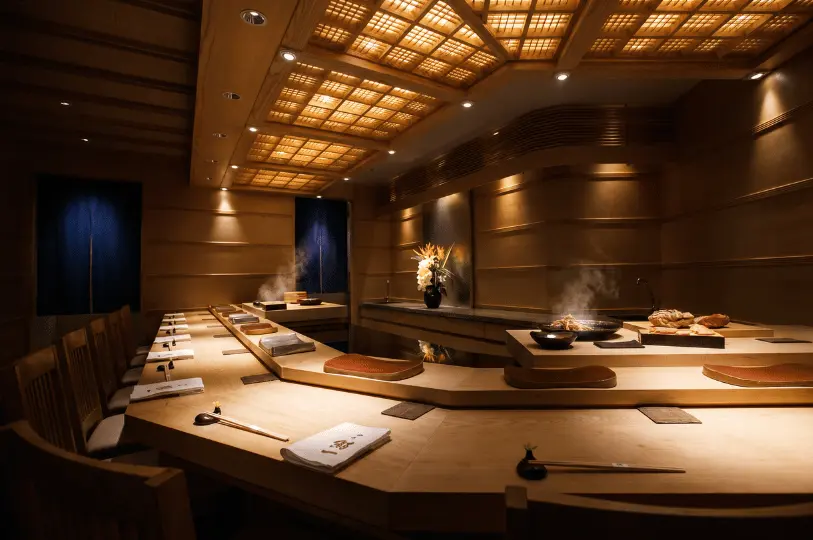 An intimate, upscale Japanese sushi counter with minimalist wooden seating, warm indirect lighting, elegant wooden ceiling latticework, and steam rising gently behind the prep area.