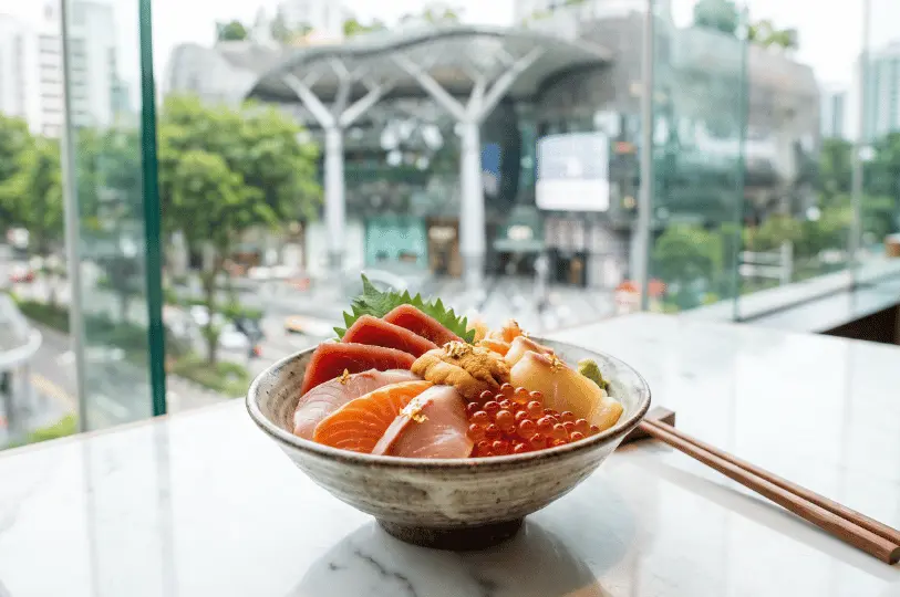 A ceramic bowl filled with a luxury chirashi don—featuring thick cuts of tuna, salmon, yellowtail, uni, and glistening ikura topped with gold leaf—set on a marble counter before a floor-to-ceiling window overlooking Orchard Road.