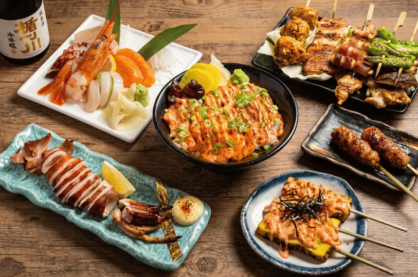 A vibrant Japanese food spread on a dark wooden table, featuring fresh sashimi, a aburi salmon don bowl, grilled whole squid, a platter of yakitori skewers, and mentaiko tamagoyaki alongside a bottle of sake.