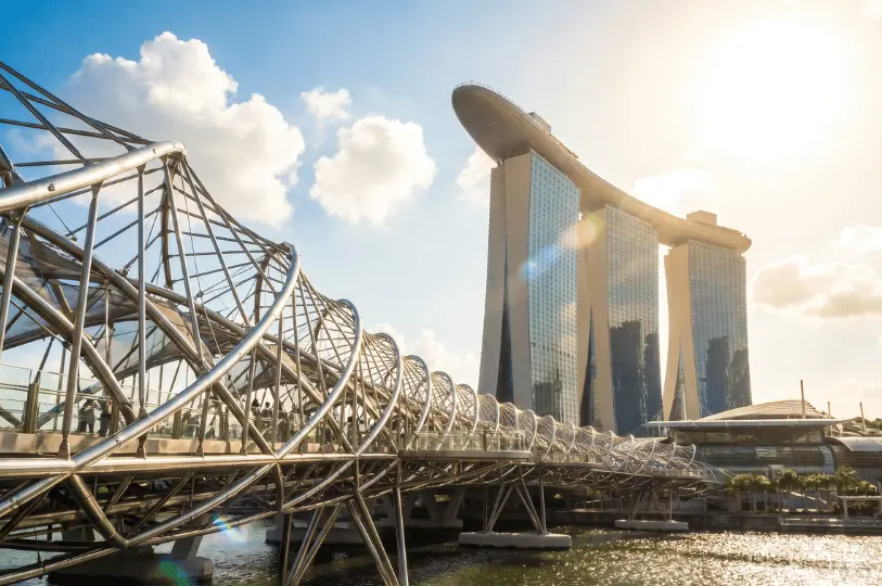 The metallic, spiral structure of the Helix Bridge stretching across the water in the foreground, with the iconic three-towered Marina Bay Sands hotel rising in the sunny background.