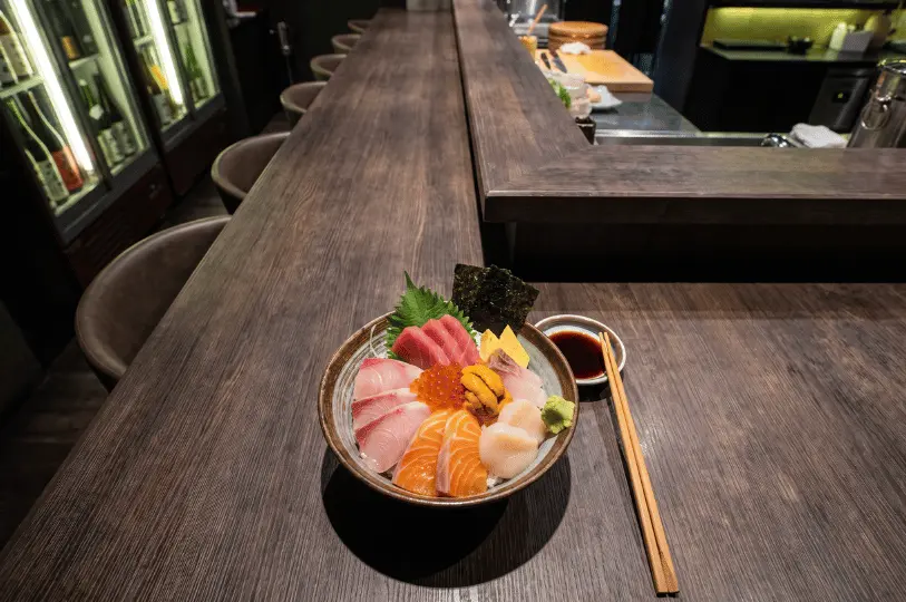 A bowl of premium kaisen don topped with assorted sashimi, sea urchin, scallops, and salmon roe resting on a dark wooden sushi bar counter, accompanied by wooden chopsticks and a small dish of soy sauce.
