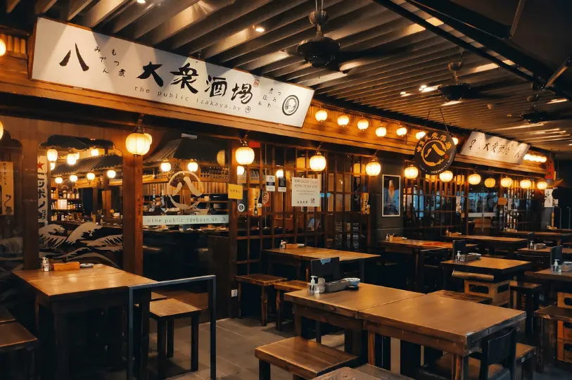 Wooden storefront and traditional outdoor seating area of The Public Izakaya by Hachi illuminated by warm hanging lanterns.