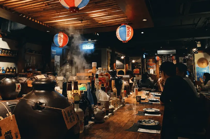 Lively counter bar seating at The Public Izakaya by Hachi, featuring large sake jars, steaming dishes, and Orion paper lanterns.