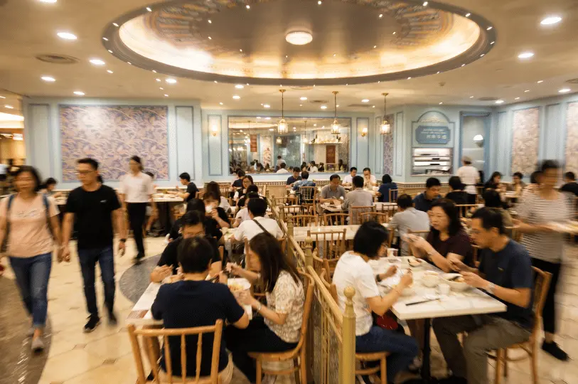 A bustling, crowded scene inside the beautifully decorated Food Opera food court, packed with diners enjoying their meals at marble tables while blurred figures walk through the aisles.