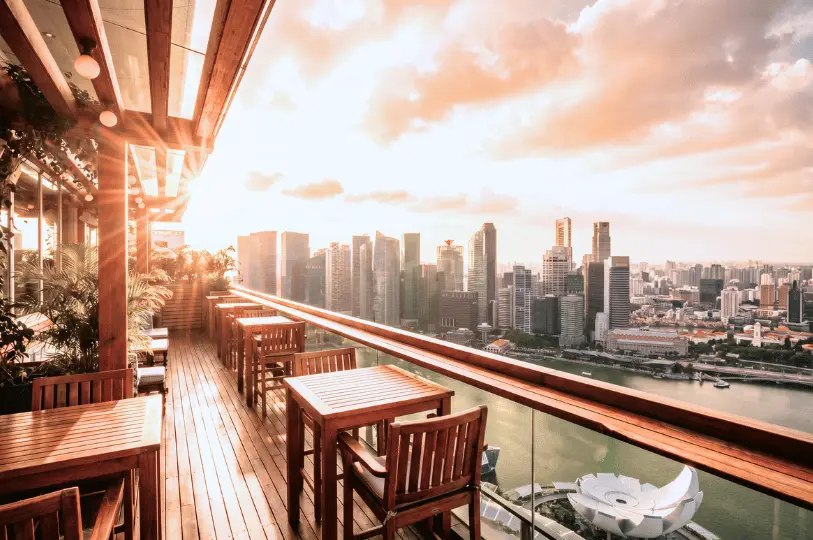 An empty outdoor dining terrace featuring wooden tables and chairs under a pergola, offering a panoramic sunset view of the Singapore city skyline and Marina Bay.