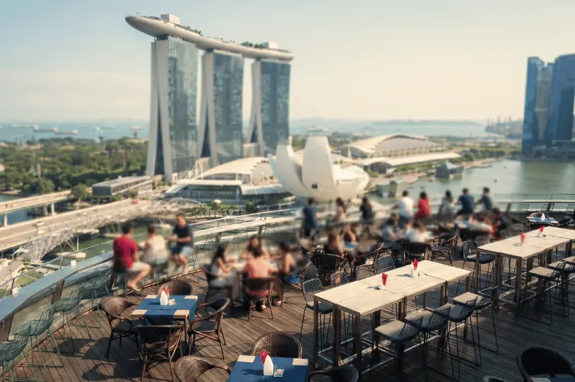 A bustling outdoor rooftop bar with guests seated at high tables, overlooking a softly blurred background of the Marina Bay Sands hotel, the ArtScience Museum, and the bay.