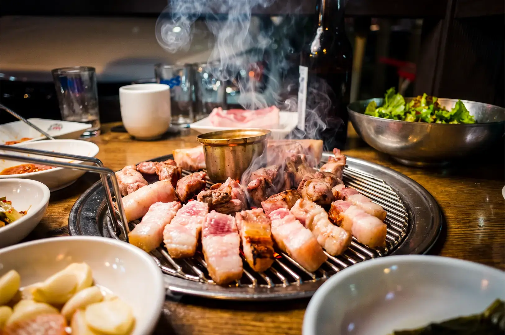 Korean BBQ close up shot with pork cuts on a griller surrounded by side dishes in bowls.