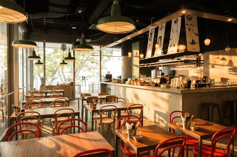 A warm, sunlit dining room at Tanuki Raw with an industrial-chic design, featuring wooden tables, red and natural wood chairs, a concrete bar counter, and large windows offering a view of lush greenery.