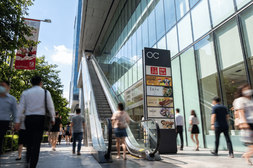 A bustling daytime street-level view outside the Orchard Central mall. A long, outdoor escalator carries people upward alongside the modern, glass-paneled facade of the building. In the foreground, blurred pedestrians walk along the sidewalk past a tall black directory sign displaying logos for mall tenants, including Uniqlo, Don Don Donki, and Joie.