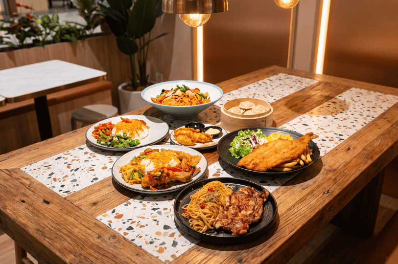 A warm, overhead-lit wooden dining table featuring terrazzo tile inlays, covered with an eclectic and abundant spread of comfort foods. The dishes include a large fried fish cutlet with fries, spaghetti topped with grilled chicken, two hearty rice plates with meat and vegetables, seafood pasta, golden fried dumplings, and a bamboo steamer basket of xiao long bao.