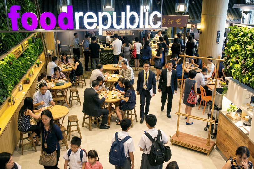 An overhead view of a highly crowded Food Republic food court during a busy lunch hour. Office workers, students, and families navigate tight aisles and share round wooden tables under a large neon sign, illustrating a bustling and overwhelming mall dining environment.