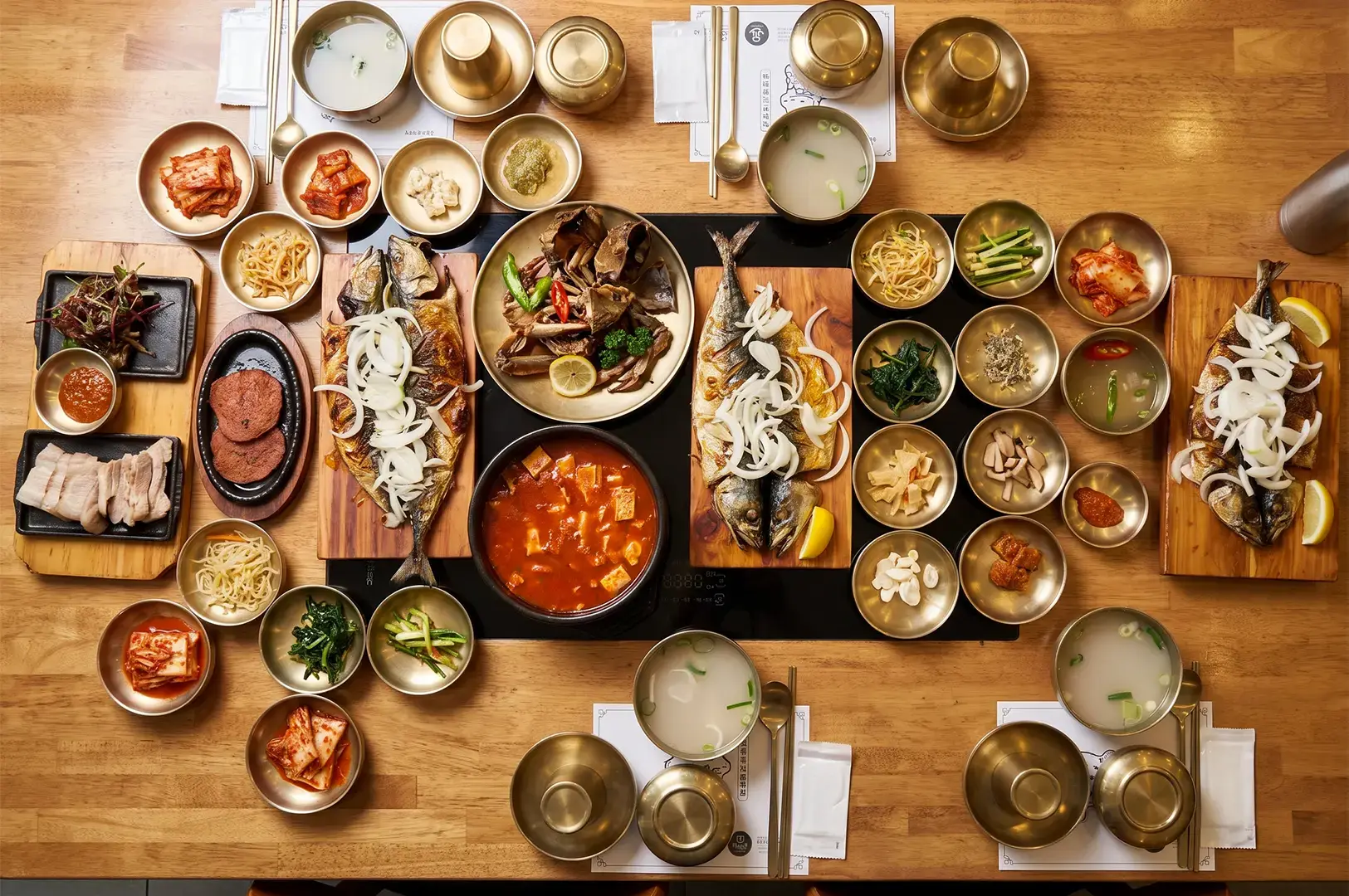 Flat lay wide shot of a Korean meal with fish, meat, and soup surrounded by a wide variety of side dishes illuminated by warm yellow lights.