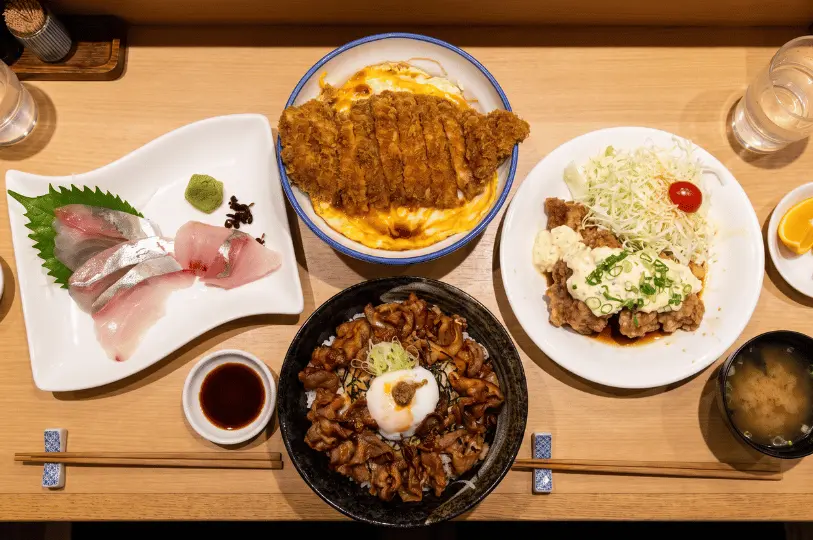 A top-down view of a hearty Japanese feast spread across a light wooden table. The meal includes a bowl of golden katsudon, a savory sliced meat and rice bowl topped with a soft-boiled egg, crispy chicken nanban with tartar sauce, a plate of fresh sashimi, miso soup, and chopsticks.